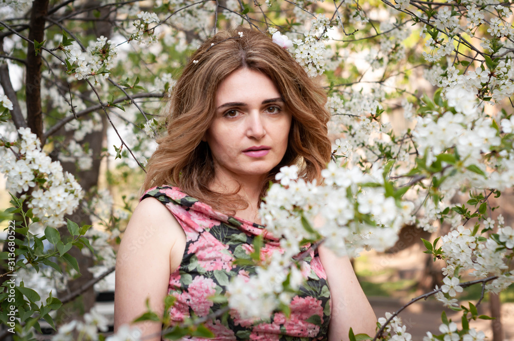 soft focus of attractive woman with long hair in pink and black dress with floral print standing near tree surrounded white blooming cherry branches and looking at camera in sunny garden soft focus of attractive woman with long hair in pink and black dress with floral print standing near tree surrounded white blooming cherry branches and looking at camera in sunny garden