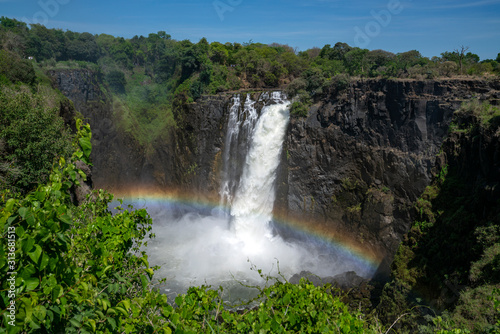 Large stream of water is flowing from the Zambezi river in to the Victoria Falls  with on top a rainbow.