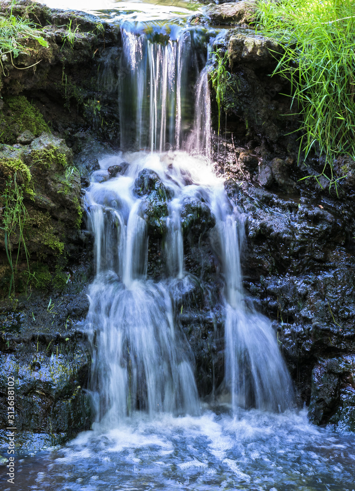 a small waterfall in a rural setting over a rocky coast with light green grass and moss on both sides