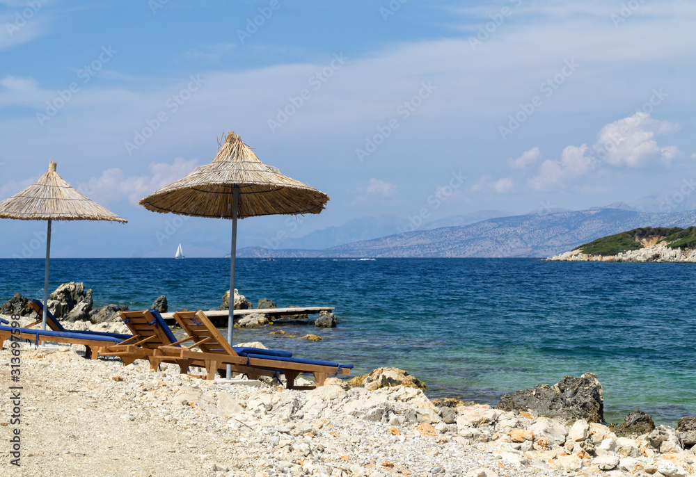 Fototapeta premium Rocky beach on the shore of the Ionian Sea with sunbeds and straw umbrellas. Blue and tourquse sea in background, nobody. Sunny Summer day. Ksamil, Albania, Europe.