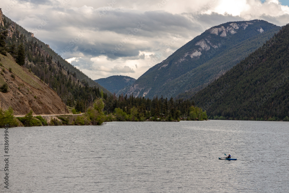 Large lake surface surrounded by tall mountains, a sole person in a kayak rowing on the water Large lake surface surrounded by tall mountains, a sole person in a kayak rowing on the water