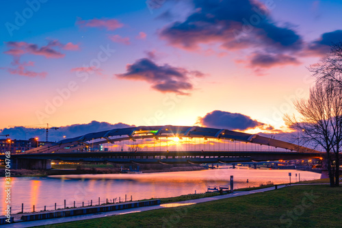 Suspension bridge Kotlarski over the Vistula River in Cracow (Krakow), Poland, against purple and orange sunset light with water reflelection. Outstanding sun lens flare.