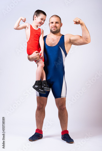 Men in wrestling tights  holds the boy with one hand and they both show bicepson a white isolated background. Dad and son have been fooling around forever.