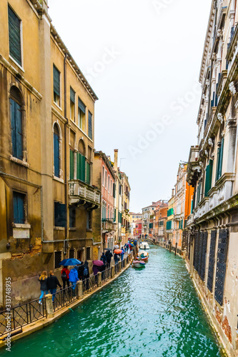 Rainy day in Venice city, Italy. Narrow water canal between colorful Venetian gothic architecture buildings with bridge connecting the islands. People/ Tourists walking with umbrellas; boats docked.