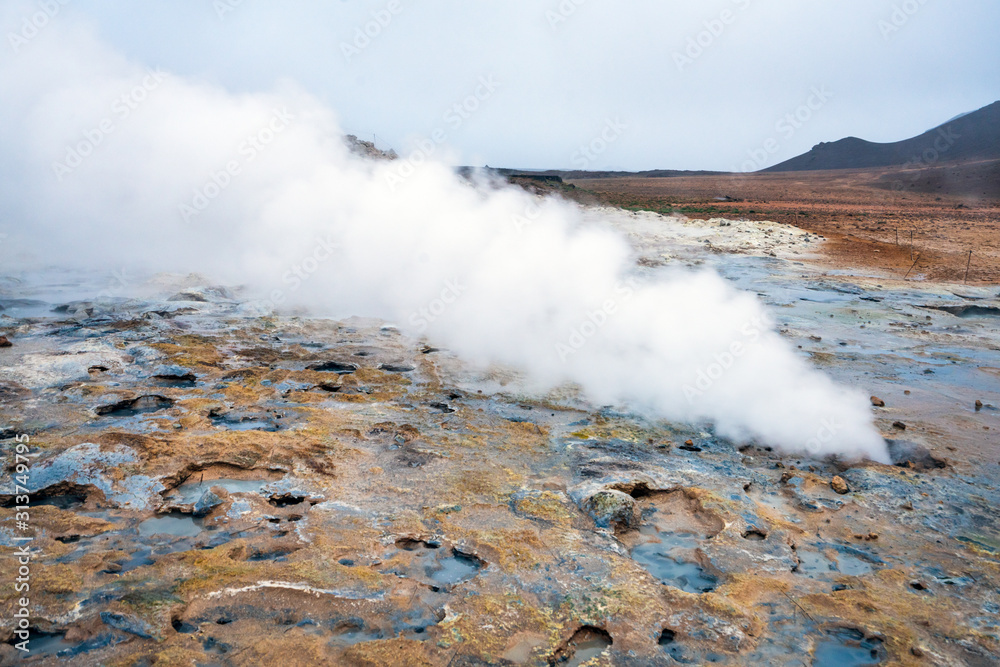 Hot sulfuric steam vent spewing sulphur steam in the hot sulfuric and geothermal area of Namaskard in Myvatn/Iceland. Color and mineral rich textured muddy ground infront.