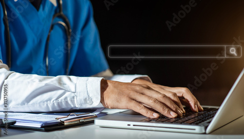 Male doctor hands typing on laptop computer keyboard,searching medical information with textbook on the desk at office. Online medical,medic tech, emr, ehr concept.