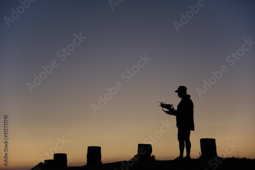 A man (with his drone) silhouette is standing on a hilltop with gold blue gradient sky in an evening