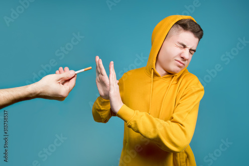 a teenager in a yellow sweatshirt on a blue background shows disgust when they hold out a cigarette, he tries to push the offer away with his hands photo