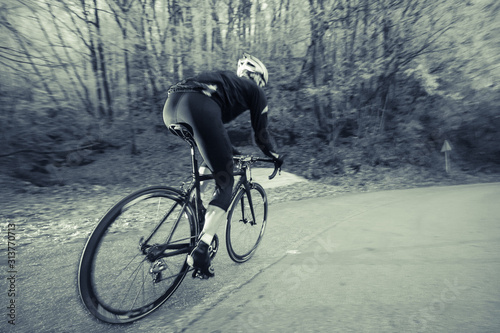 A road cyclist on a black bike is riding in a cold environment. Trees with white patches of snow are seen in the background. Black and white edited image.