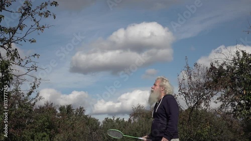 Sportive senior Caucasian man with splendid grey hair and beard is playing badminton. Beautiful portrait of active European people in summer leisure. Outdoor lifestyle scene in slow motion footage.