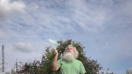 Sportive senior Caucasian man with splendid grey hair and beard is playing badminton. Beautiful portrait of active European people in summer leisure. Outdoor lifestyle scene in slow motion footage.