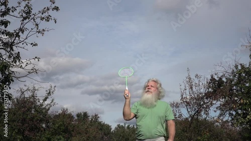 Sportive senior Caucasian man with splendid grey hair and beard is playing badminton. Beautiful portrait of active European people in summer leisure. Outdoor lifestyle scene in slow motion footage.