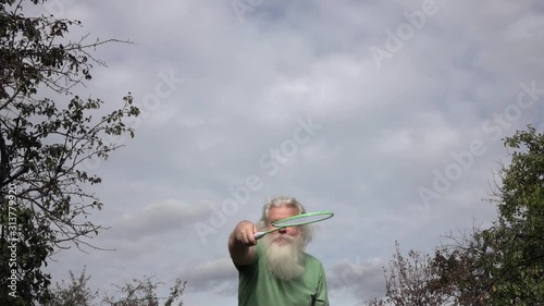 Sportive senior Caucasian man with splendid grey hair and beard is playing badminton. Beautiful portrait of active handsome European people in summer leisure. Outdoor lifestyle scene in slow motion