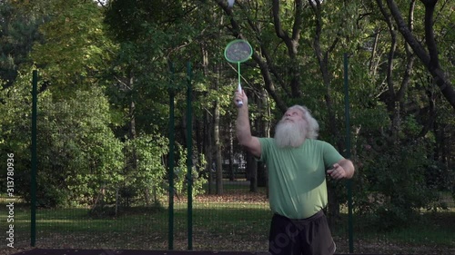Sportive senior Caucasian man with splendid grey hair and beard is playing badminton. Beautiful portrait of active handsome European people in summer leisure. Outdoor lifestyle scene in slow motion