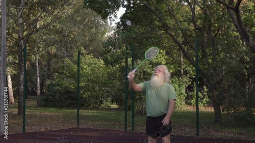Sportive senior Caucasian man with splendid grey hair and beard is playing badminton. Beautiful portrait of active handsome European people in summer leisure. Outdoor lifestyle scene in low sunshine.