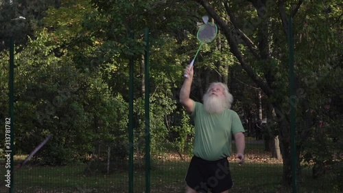 Sportive senior Caucasian man with splendid grey hair and beard is playing badminton. Beautiful portrait of active handsome European people in summer leisure. Outdoor lifestyle scene in low sunshine.