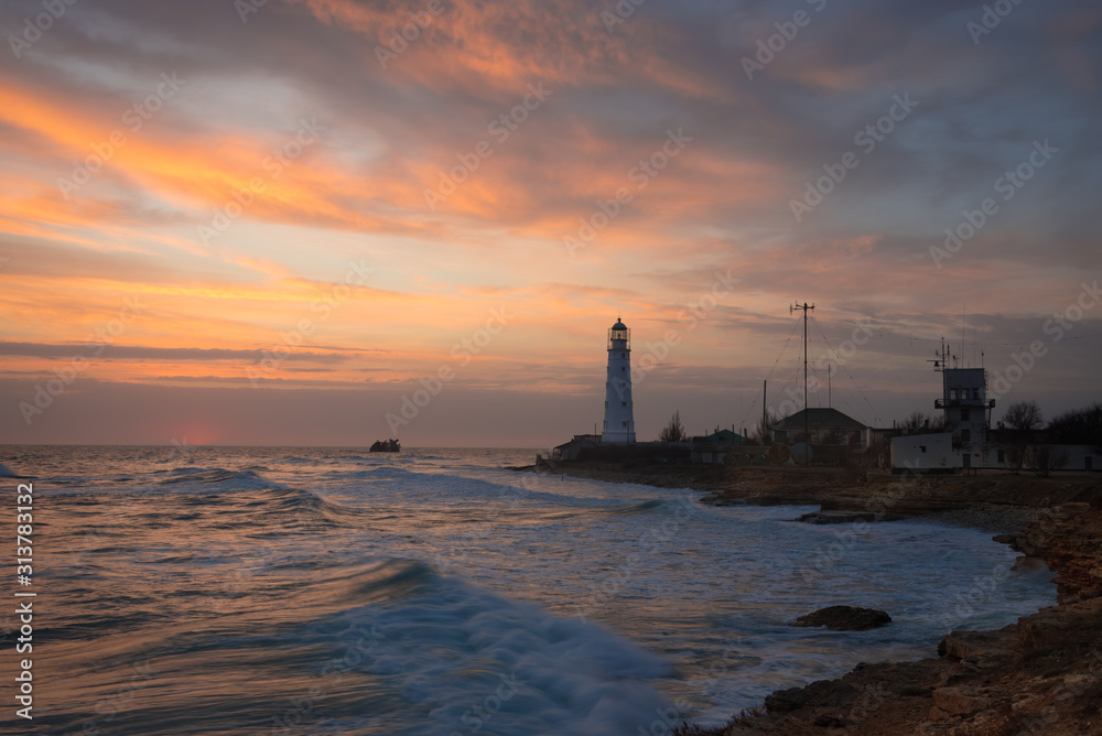 Orange sunset on the Cape with the picturesque Tarkhankut lighthouse, the sea coast of Crimea and a sunken ship in the background