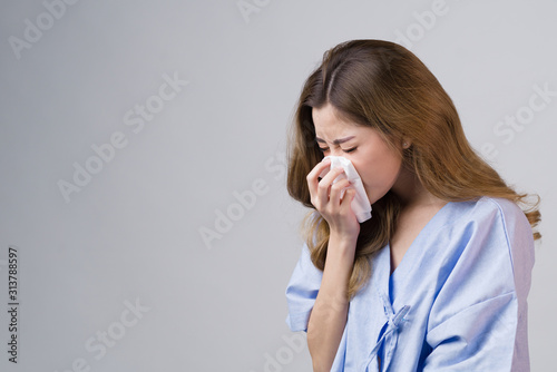 Portrait of a beautiful Asian woman in patient gown hold handkerchief to blow her nose, flu cold sneezing isolated in gray background with copy space, concept healthcare insurance, hayfever allergy.