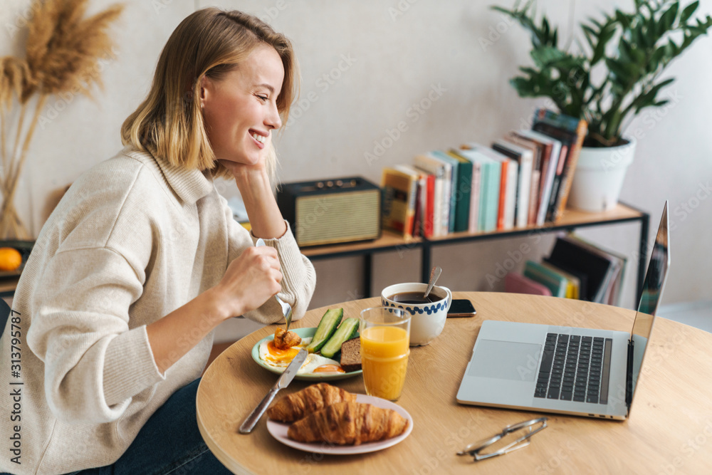 Photo of happy blonde woman using laptop while having breakfast