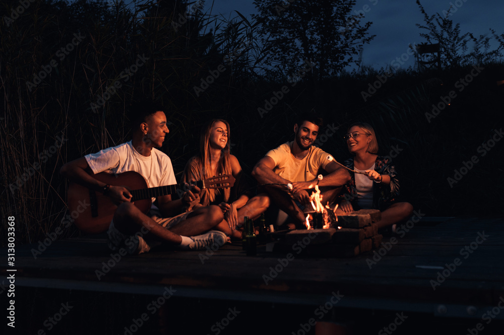 Group of young friends enjoying at the lake at night. They sitting around the fire singing and having fun at camping. Group of young friends enjoying at the lake at night. They sitting around the fire singing and having fun at camping.