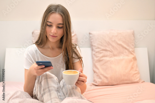 Young Woman Sitting In Bed With Smartphone And Cup of Coffee