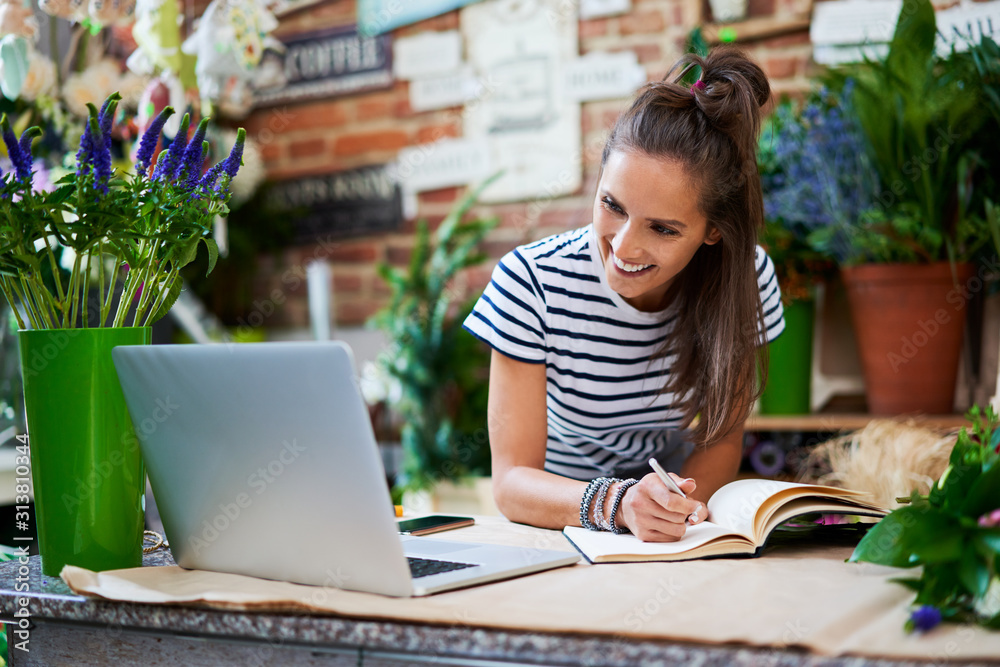 Joyful florist looking at laptop with client orders and writing them down in notebook Joyful florist looking at laptop with client orders and writing them down in notebook