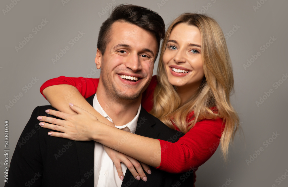 Elegant couple. Close up photo of a charming blonde girl in a red dress, who is hugging her boyfriend in a suit from the back, looking in the camera and smiling. Elegant couple. Close up photo of a charming blonde girl in a red dress, who is hugging her boyfriend in a suit from the back, looking in the camera and smiling.