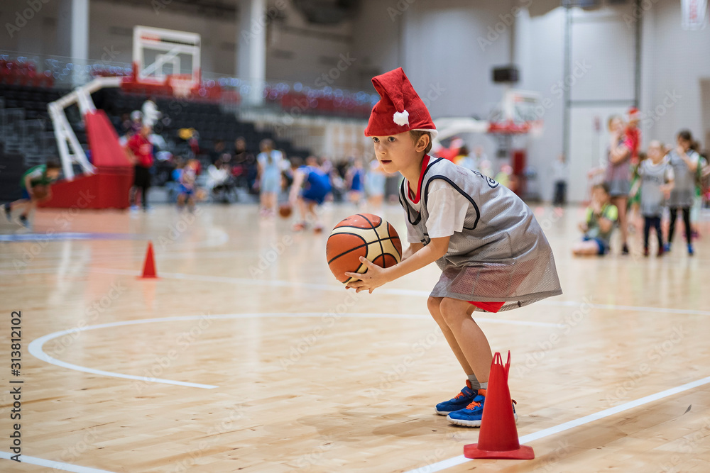 Fototapeta premium Young boy with Santa Clause hat exercises with a basketball ball in a sports hall