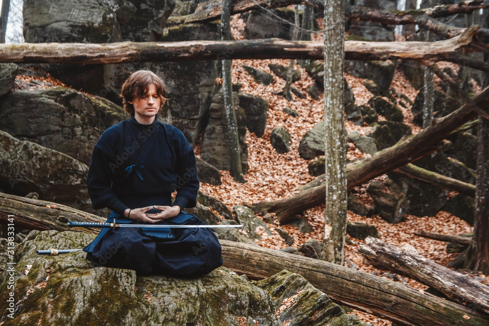 Young man in black kimono with a sword meditates and concentrates on rocks and forest background. Place for text or advertising Young man in black kimono with a sword meditates and concentrates on rocks and forest background. Place for text or advertising