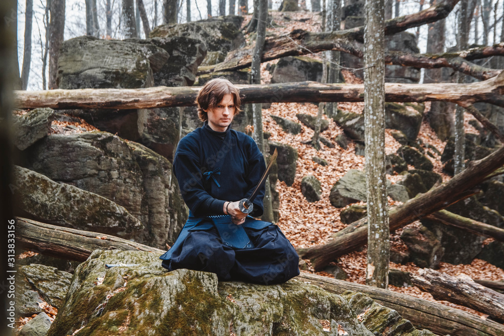 Young man in black kimono with a sword meditates and concentrates on rocks and forest background. Place for text or advertising Young man in black kimono with a sword meditates and concentrates on rocks and forest background. Place for text or advertising