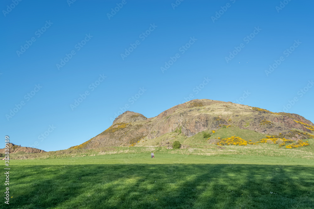 View of Salisbury crags on Holyrood Park with green spaces, tourists walking and looking, blue sky on background, in Edinburgh