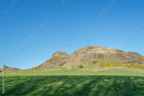 View of Salisbury crags on Holyrood Park with green spaces, tourists walking and looking, blue sky on background, in Edinburgh