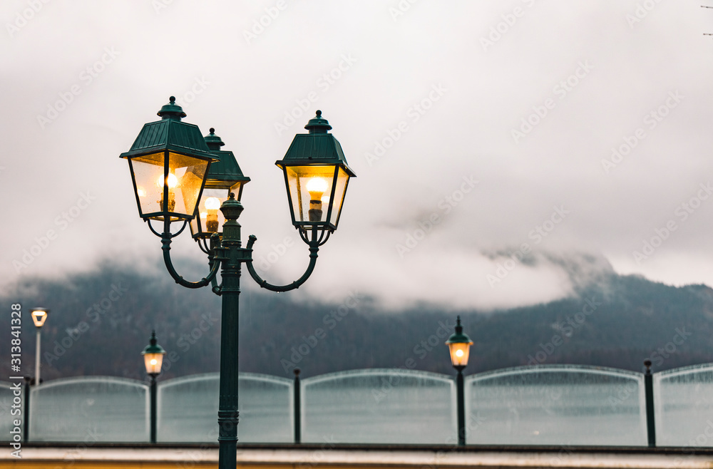 moody foggy weather time harbor area with street lantern yellow lighting on waterfront district foreground and picturesque gorgeous mountain landscape background in clouds, copy space