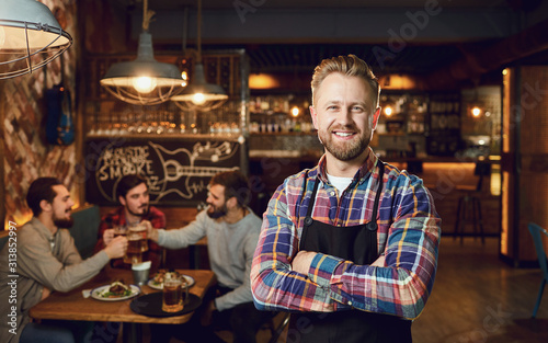 Bearded waiter barista bartender standing against the background of the pub bar