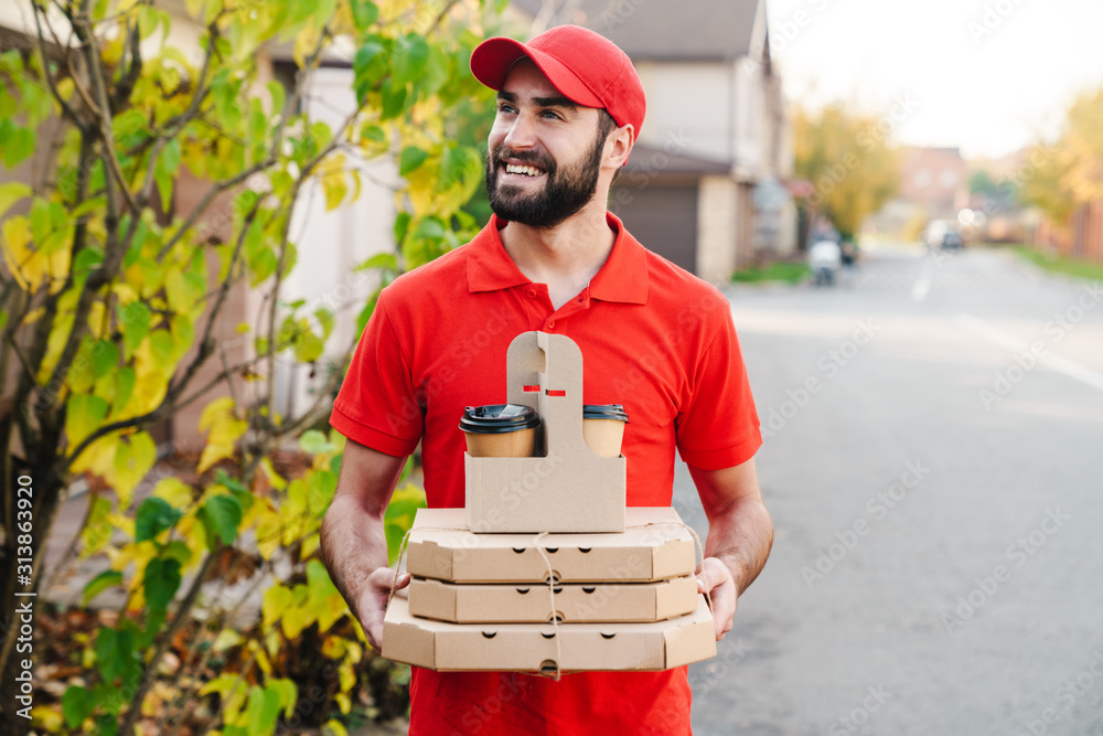 Image of smiling young delivery man holding pizza boxes and coffee