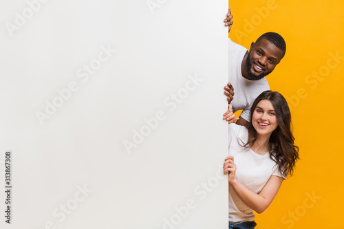 Joyful interracial couple peeking from behind white advertisement board photo