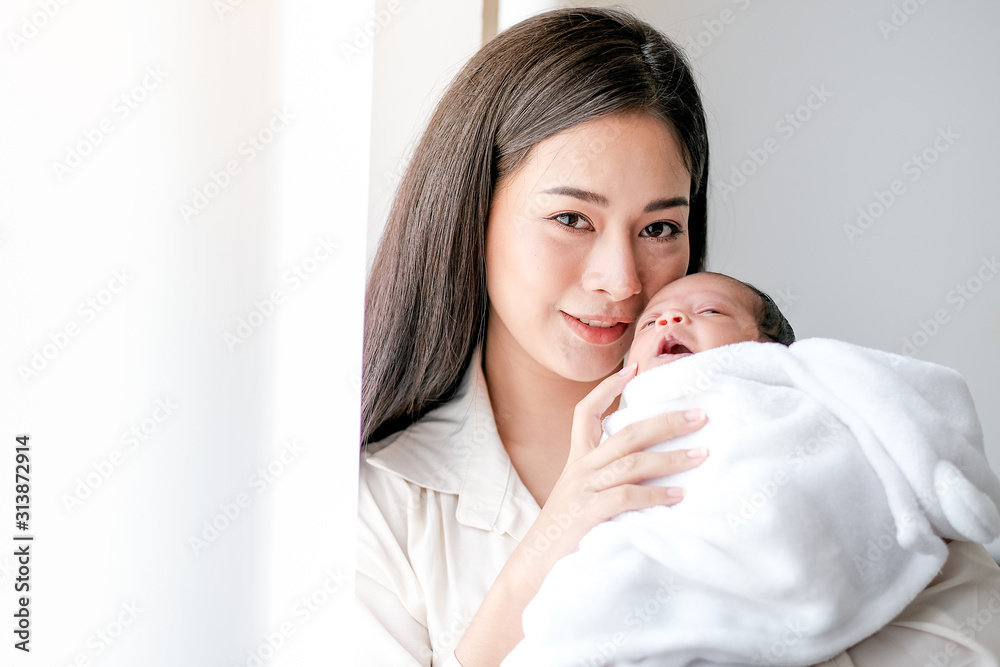 Portrait of young mother hold newborn baby look to camera and stand in front of white curtain with morning light. Portrait of young mother hold newborn baby look to camera and stand in front of white curtain with morning light.
