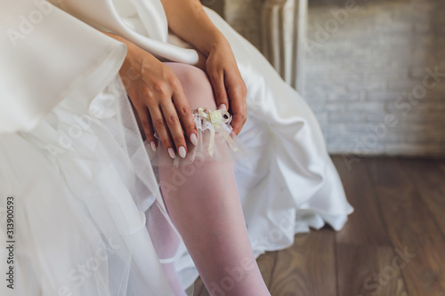 In the morning, the bride in stockings and a white wedding dress wears a garter on her leg, the bride is holding her hands for the garter. photo