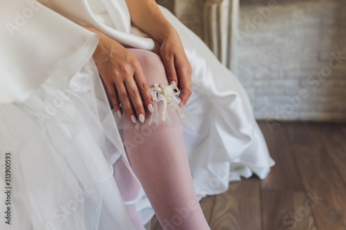 In the morning, the bride in stockings and a white wedding dress wears a garter on her leg, the bride is holding her hands for the garter.