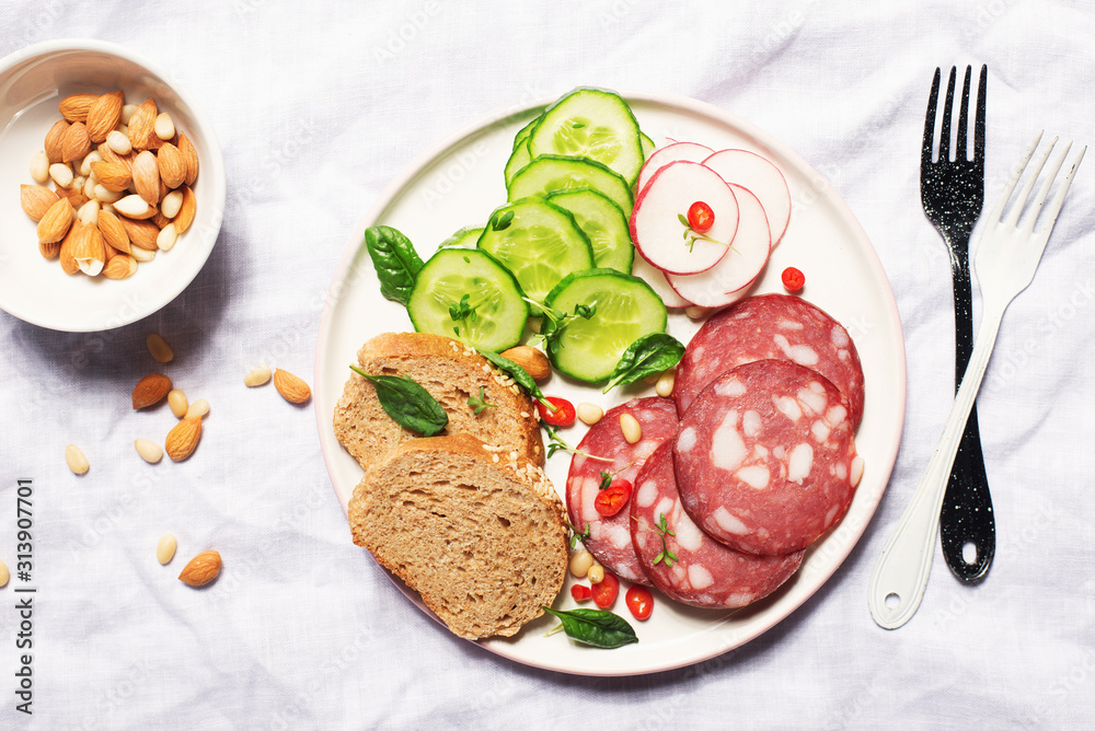 Appetizer with fresh vegetables cucumber, radishes, spinach, micro-greens, and slices of salami on a dark plate in hard bright light. Trend snapshot. Top view. Hard shadows