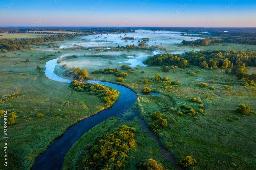 An amazing aerial view of sunrise over foggy river. River is surrounded by green fields and forest. Amazing landscape