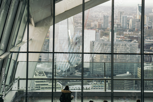 Young woman with green jacket looking at the panoramic view from the viewpoint of the London Sky Garden building. She is drinking hot coffee