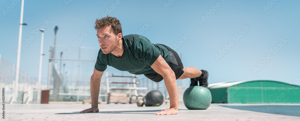 Fitness man banner panorama. Athlete strength training pushup balancing legs on medicine ball for advanced core body workout push-ups floor exercises at outdoor gym. - obrazy, fototapety, plakaty 