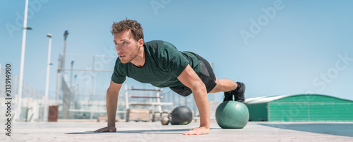 Fitness man banner panorama. Athlete strength training pushup balancing legs on medicine ball for advanced core body workout push-ups floor exercises at outdoor gym.