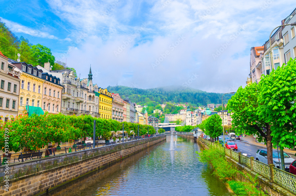 Karlovy Vary (Carlsbad) historical city centre with Tepla river central embankment, colorful beautiful buildings, Slavkov Forest hills with green trees background, West Bohemia, Czech Republic