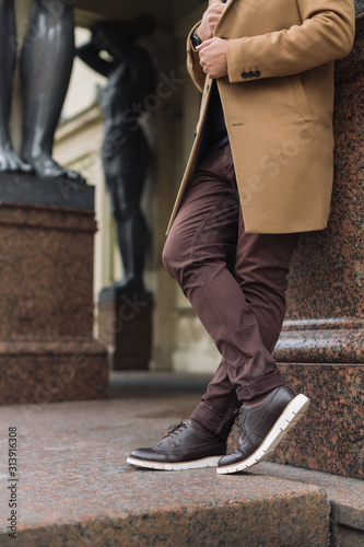 Cropped portrait of unrecognizable handsome man in elegant autumn beige coat, brown pants and dark leather shoes with white soles. Young stylish hipster is leaning against the wall. Fashion concept.
