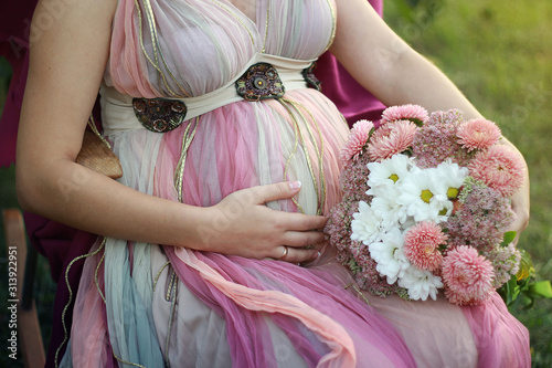  Pregnant woman in a beautiful powdery dress sitting on an armchair with a bouquet of beautiful flowers on a summer evening