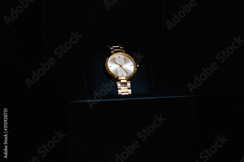 Men's golden colored, classic round wrist watch with metal strap in an open black box in a dark room. Isolated on black background. Accessories in an open gift package.