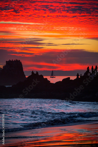 The sunset colors the sky brilliant colors people watch a distant sail boat from the rocks at Crescent Cove  near Laguna Beach  California.
