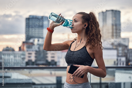 Side shot of young athletic woman drinking water from sport bottle while listening to music on city background